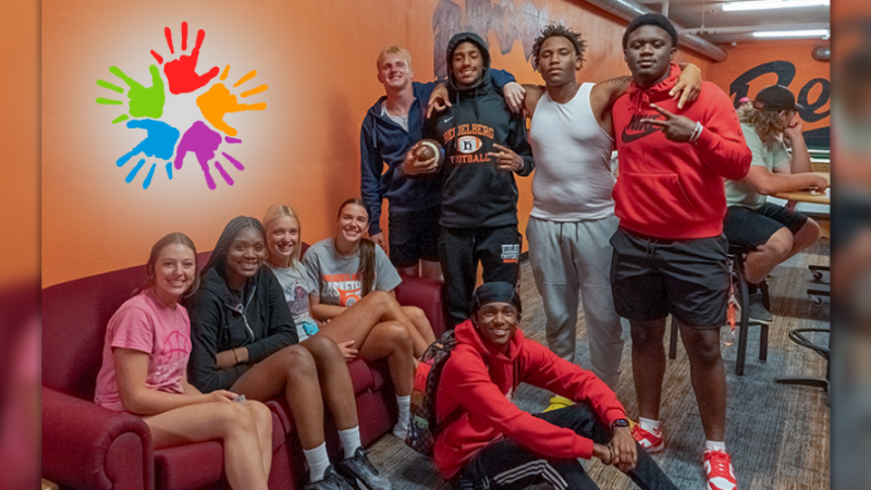 A collection of several diverse students smiling in the Brown hall basement lounge. The wall is orange with an image of five mulitcolor hands superimposed. 