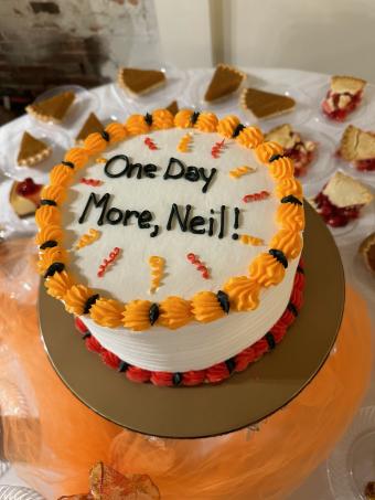 A white cake with yellow and orange frosting details that reads "One Day More, Neil!"